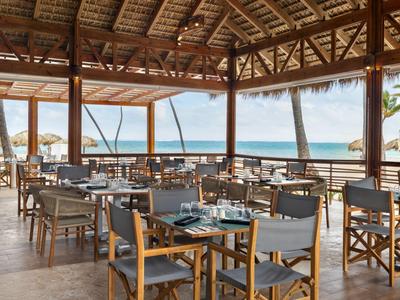 Restaurante abierto con sillas y mesas de madera con vista al mar en día despejado.