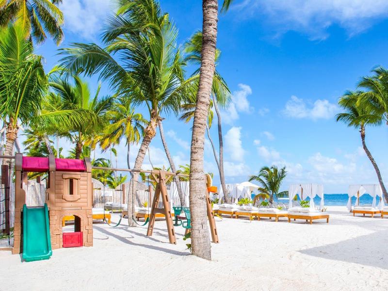 Área de juegos con columpios y estructura para trepar en la playa con palmeras y tumbonas bajo cielo azul