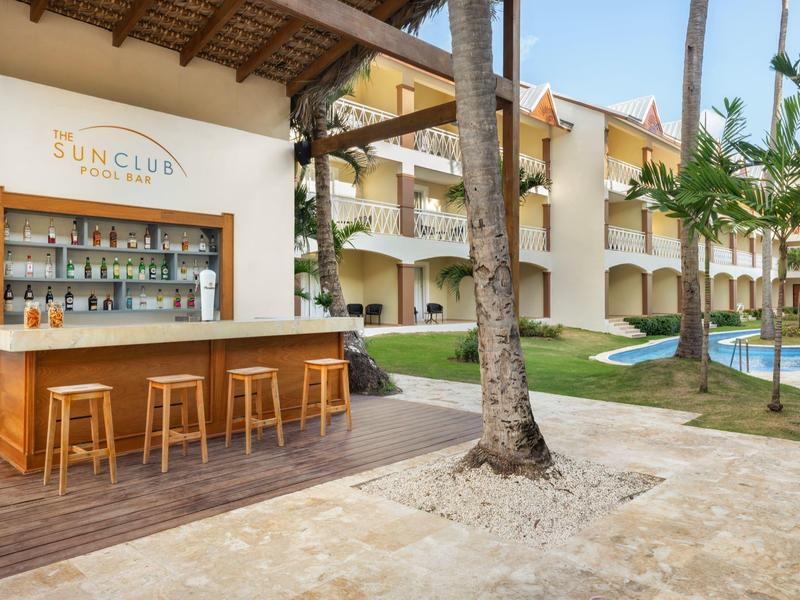 Área de bar al aire libre con taburetes de madera junto a piscina y edificio hotelero en entorno tropical.