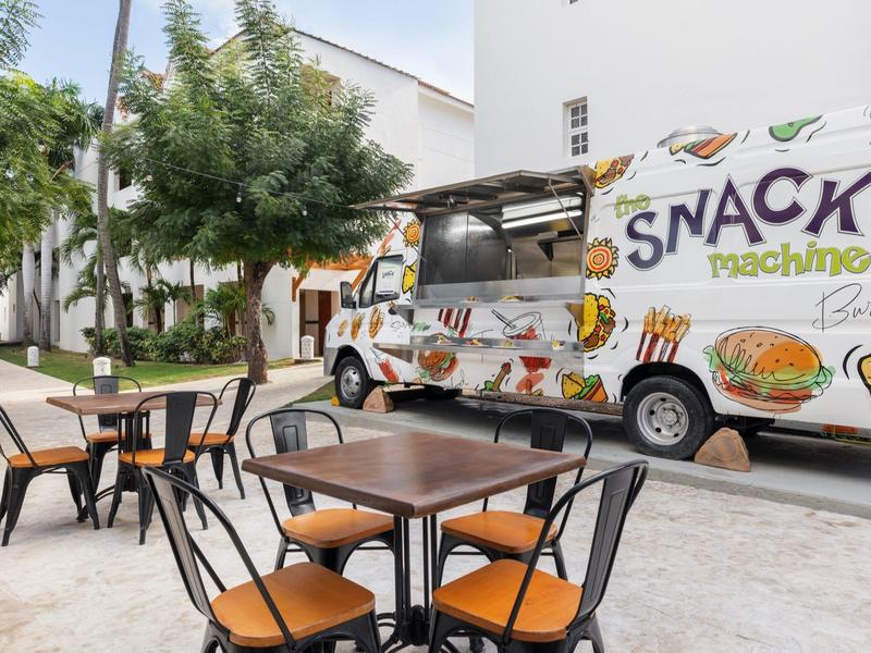 Área exterior de un camión de comida con mesas, sillas y árboles al fondo.