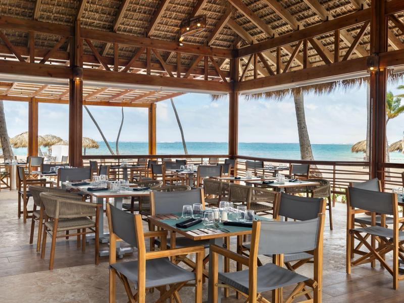Restaurante abierto con sillas y mesas de madera con vista al mar en día despejado.