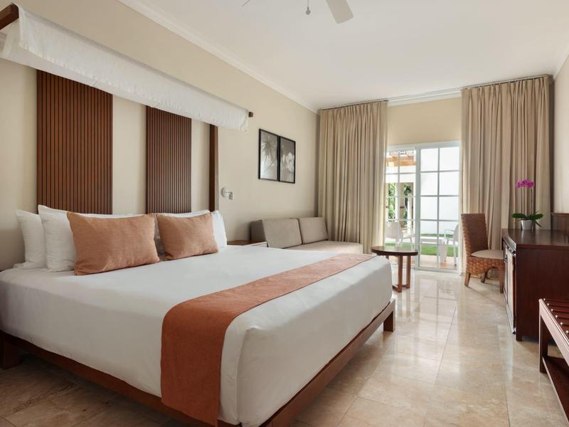 Habitación de hotel luminosa con cama grande, zona de estar y escritorio con TV, iluminada por puerta a terraza.