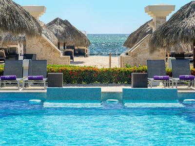 Zwembad met ligbedden en rieten daken, uitzicht op zee op de achtergrond.