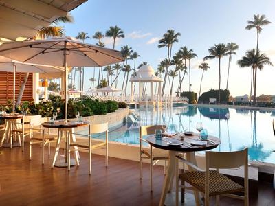 Terrasse eines Hotels mit Tischen, Stühlen und Sonnenschirmen neben einem Pool und Palmen.