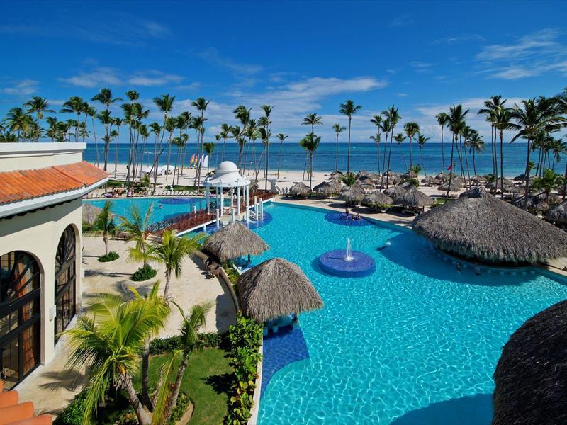 Groot hotelzwembad met palmbomen, parasols en zeezicht onder een blauwe lucht.