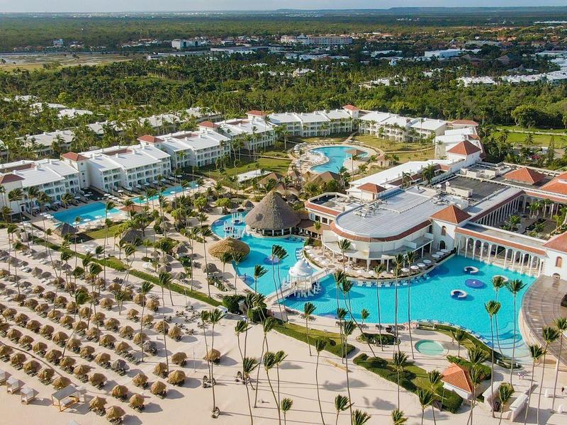 Luchtfoto van een luxe strandresort met meerdere zwembaden, parasols en een groene omgeving.