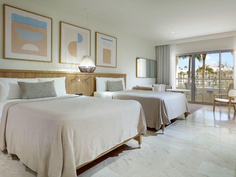 Moderne hotelkamer met twee tweepersoonsbedden, lichte muren en uitzicht op palmbomen door grote ramen.