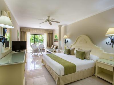 Helles Hotelzimmer mit Doppelbett, grün-weißer Bettwäsche, TV, Deckenventilator und Balkon mit Blick ins Grüne.