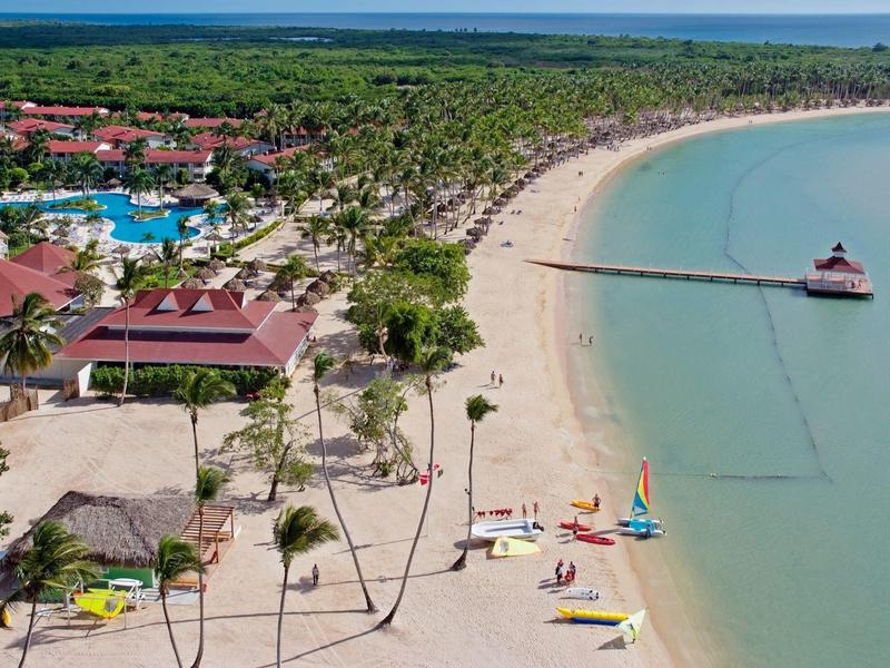 Plaża z czystą wodą, żaglówkami i pomostem otoczonym palmami i budynkami.