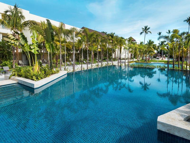 Groot zwembad omgeven door palmbomen en een meer verdiepingen tellend hotel onder een blauwe lucht.