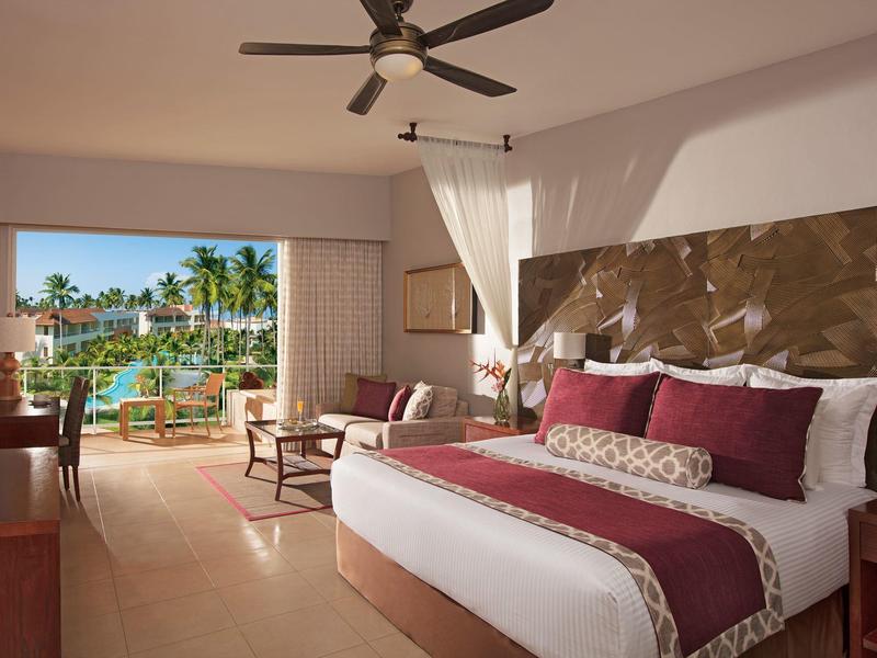 Lichte hotelkamer met groot bed, plafondventilator en uitzicht op tropisch zwembad en palmbomen.