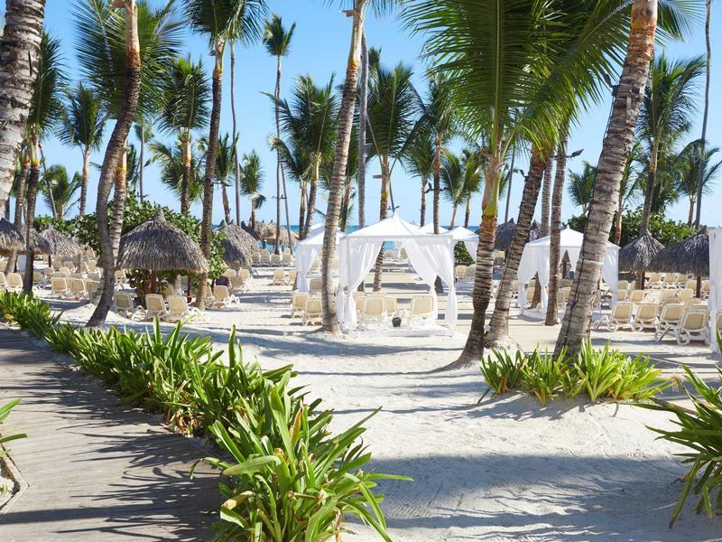 Playa con palmeras, arena y un dosel blanco con sillas para los invitados.