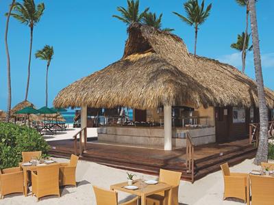 Strandbar met rieten dak, omgeven door palmbomen en zitgedeeltes in het zand.