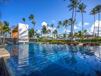 Großer Hotelpool mit blauer Wasseroberfläche, Palmen und Sonnenliegen bei klarem Himmel.