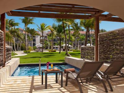 Blick von einer schattigen Terrasse mit Pool auf einen tropischen Garten und Hotelgebäude.