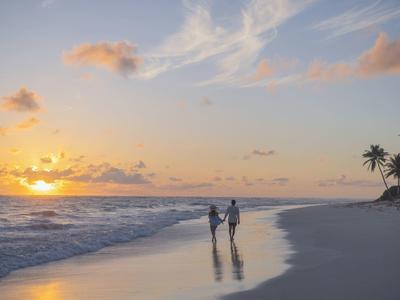 A couple holding hands walks on the beach at sunset, palm trees in the background.