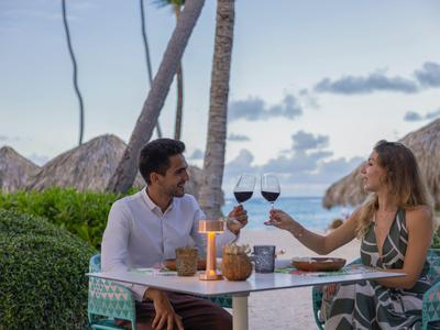 Couple drinks red wine at romantic beachside dinner with palm trees and ocean view.