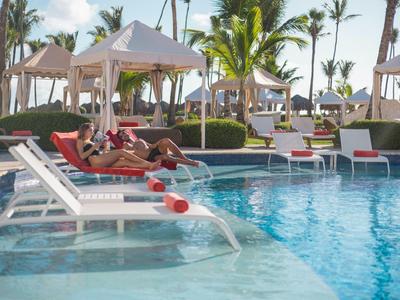 Relaxed guests lie on sunbeds by the pool with palm trees and cabanas in view.