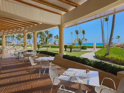 Overdekt terras met witte tafels en stoelen, uitzicht op palmbomen en zee.