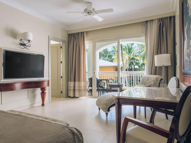 Bright hotel room with TV, seating area, balcony overlooking palm trees, and comfortable furniture.