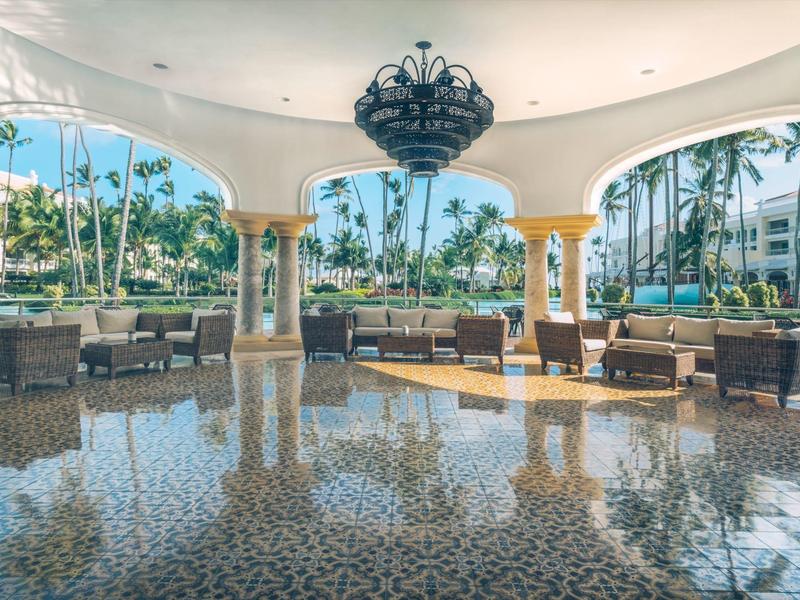 Open, elegant hotel lounge area with wicker furniture and large columns, palm trees in the background.