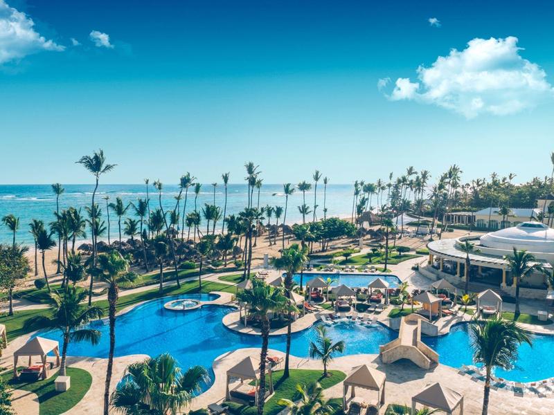Overview of a hotel resort with curved pool, palm trees, and ocean view under a blue sky.