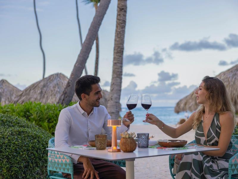 Couple drinks red wine at romantic beachside dinner with palm trees and ocean view.