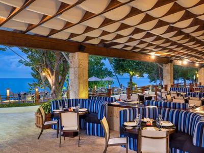 Restaurantterrasse mit blau-weiß gestreiften Sesseln, Holztischen und Blick aufs Meer bei Abenddämmerung.