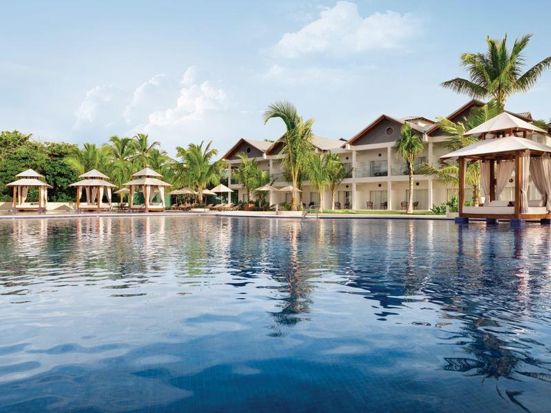 Duży basen przed ośrodkiem z altankami i palmami pod błękitnym niebem.