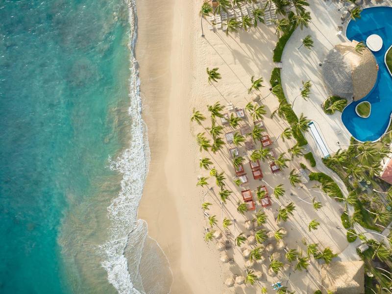 Luftaufnahme von blauem Meer, hellem Sandstrand, Palmen und einem blauen Pool mit angrenzendem Gebäude.