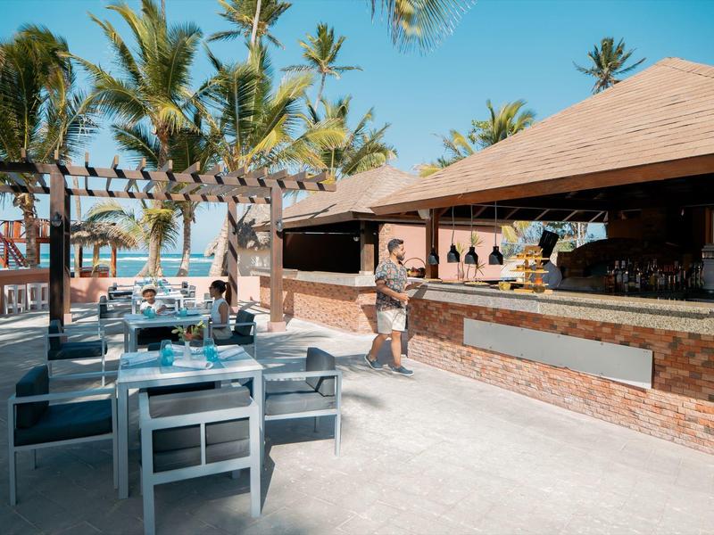 Offenes Strandrestaurant mit Holzdach, Sitzgruppen und Palmen unter blauem Himmel.