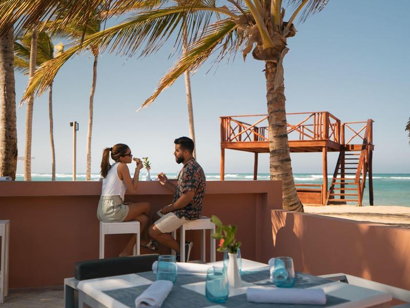 Zwei Personen sitzen an einer Strandbar aus rosa Stein, umgeben von Palmen und blauem Himmel.