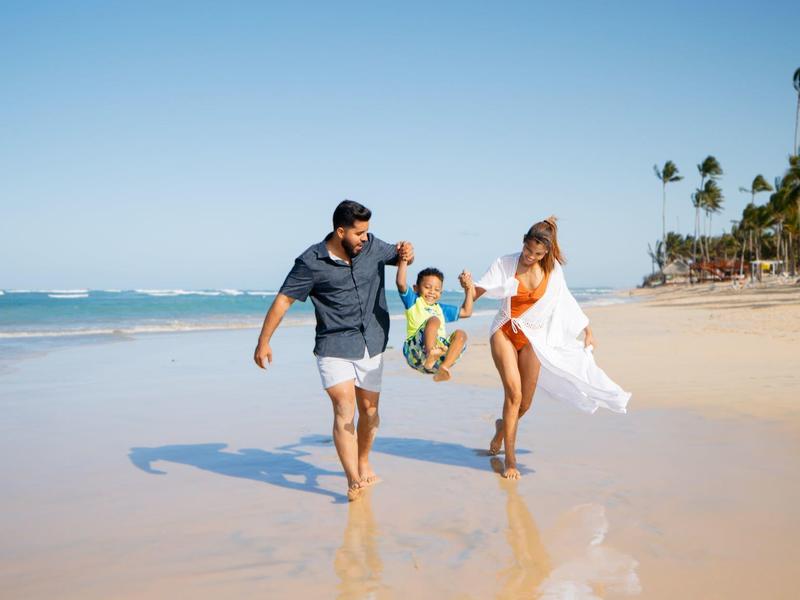 Familie läuft barfuß am Strand, Eltern halten lachendes Kind an den Händen im Sonnenschein.