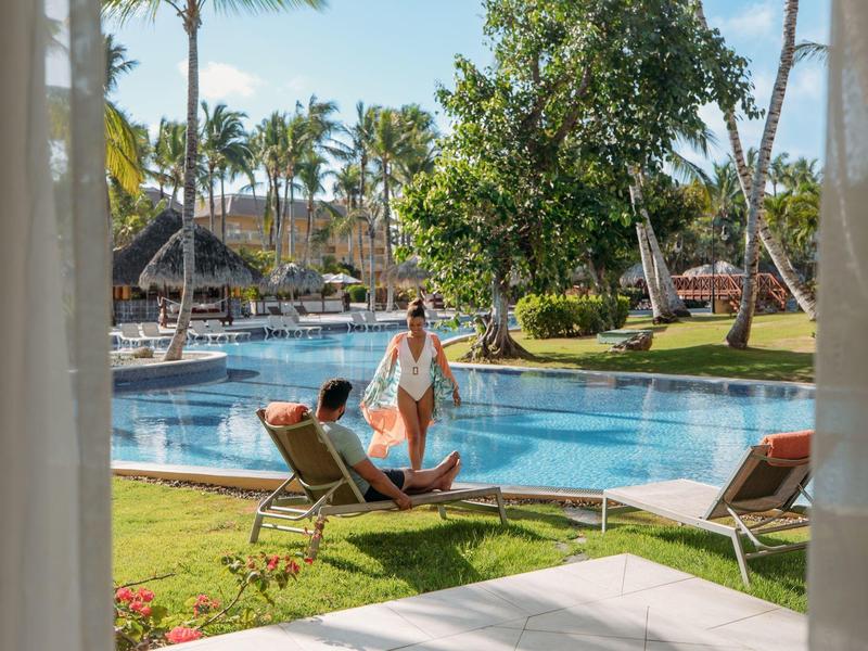 Frau neben Pool auf Liegestuhl, Palmen und Hütten im sonnigen, tropischen Resort.