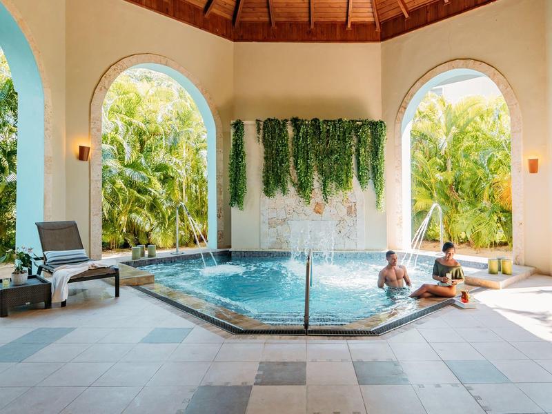 Überdachter Außenpool mit drei großen Rundbögen, Palmen, ruhiger Atmosphäre und Menschen am Wasser.