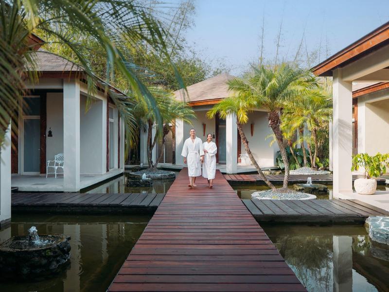 Zwei Personen gehen auf einem roten Holzsteg zwischen modernen weißen Gebäuden mit Palmen und Wasser.