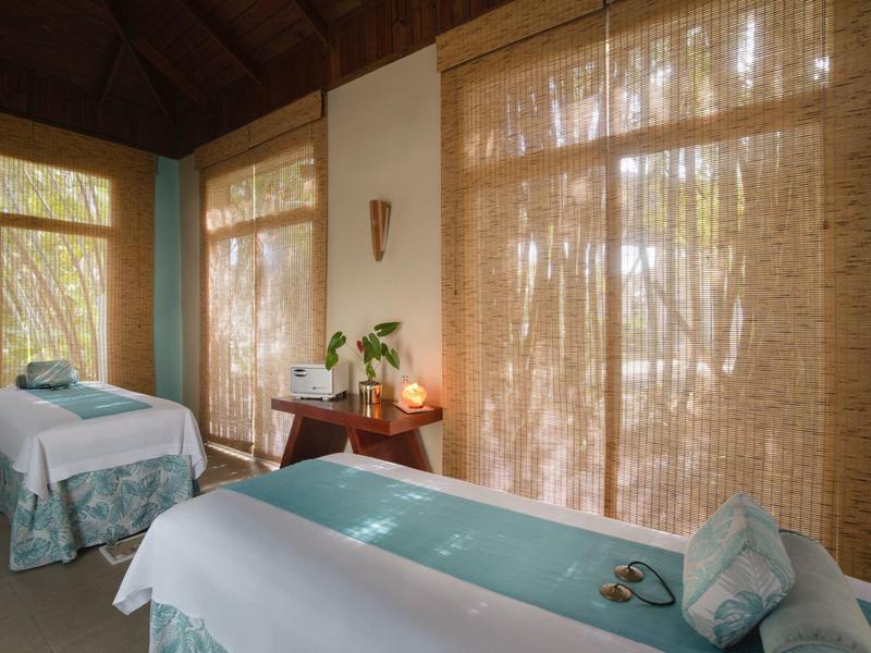 Helles Spa-Zimmer mit zwei Massageliegen, Bambusrollos und beruhigender Naturkulisse draußen.