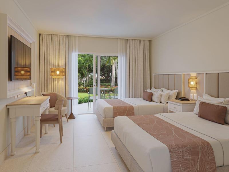 Helles Hotelzimmer mit zwei Doppelbetten, Schreibtisch, Stuhl und Blick in einen Gartenbereich.