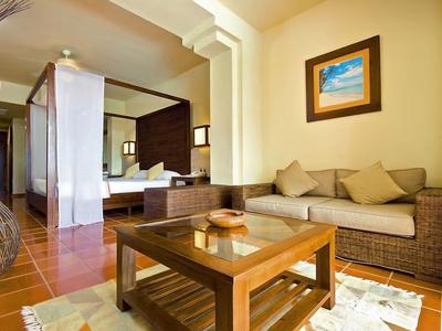 Gezellige hotel woonkamer met rotan bank, glazen salontafel en ingelijste kunst aan gele muur.