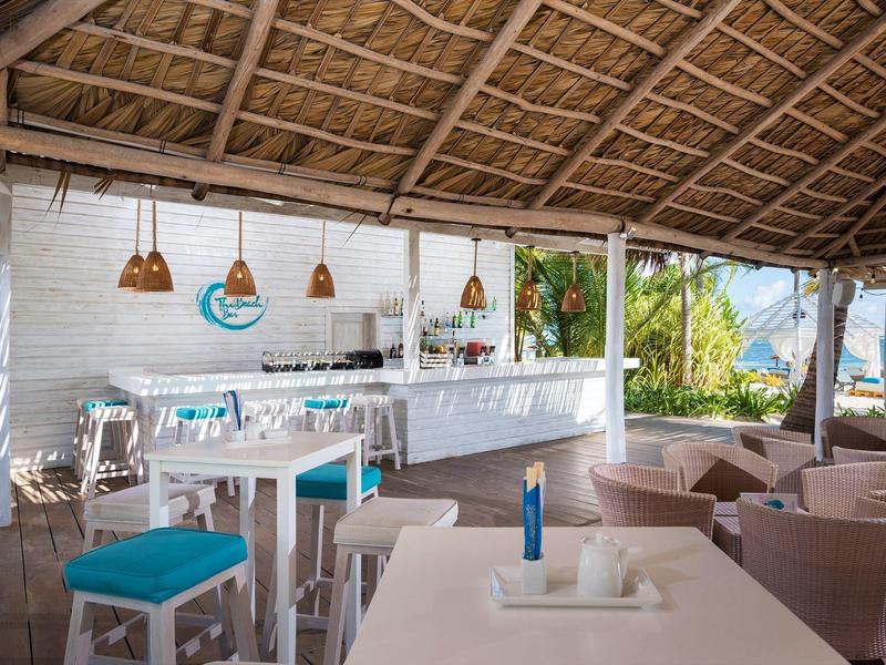 Overdekte open zitruimte met bar, stoelen en tafels omgeven door tropische begroeiing.