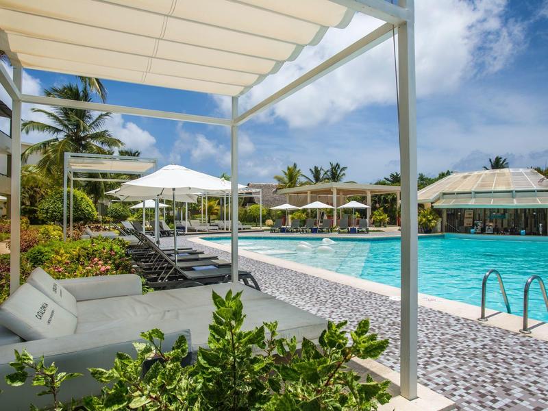 Moderne zwembadomgeving met ligstoelen, parasols en palmbomen onder blauwe lucht.