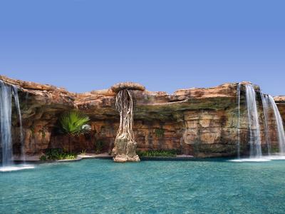 Drei Wasserfälle fließen von einem felsigen Rand in einen klaren blauen Pool unter hellem Himmel.