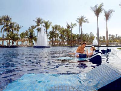 Zwei Männer entspannen auf einem Schwimmbadfloß mit einer Fontäne und Palmen im Hintergrund.