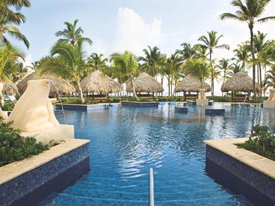 Großer Hotelpool mit Palmen und Statuen in tropischer Umgebung bei Sonnenschein.
