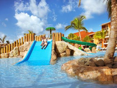 Kinderen glijden in ondiep zwembadwater in een zonnig hotelgebied met palmbomen.