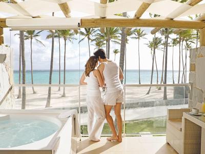 Koppel kijkt vanuit hotelkamer naar palmbomen en zee onder een helder zonnescherm.