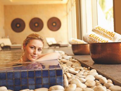 Vrouw ontspant in spa-bad met stenen vloer en decoratieve wandobjecten.