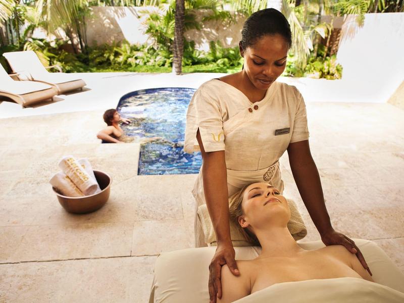 Vrouw krijgt massage in spa met relaxte zwembadgedeelte en ligstoelen op de achtergrond.