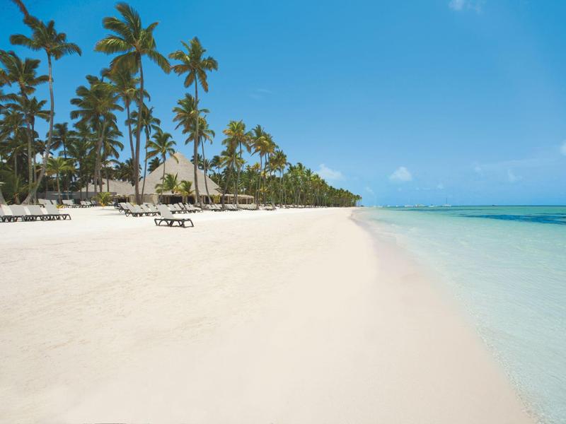 Breit wit zandstrand met palmbomen en helderblauwe zee onder een heldere lucht.