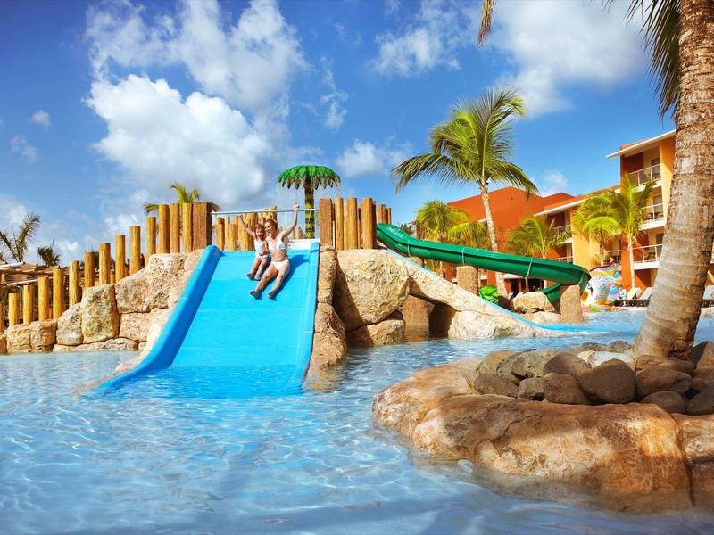 Kinderen glijden in ondiep zwembadwater in een zonnig hotelgebied met palmbomen.
